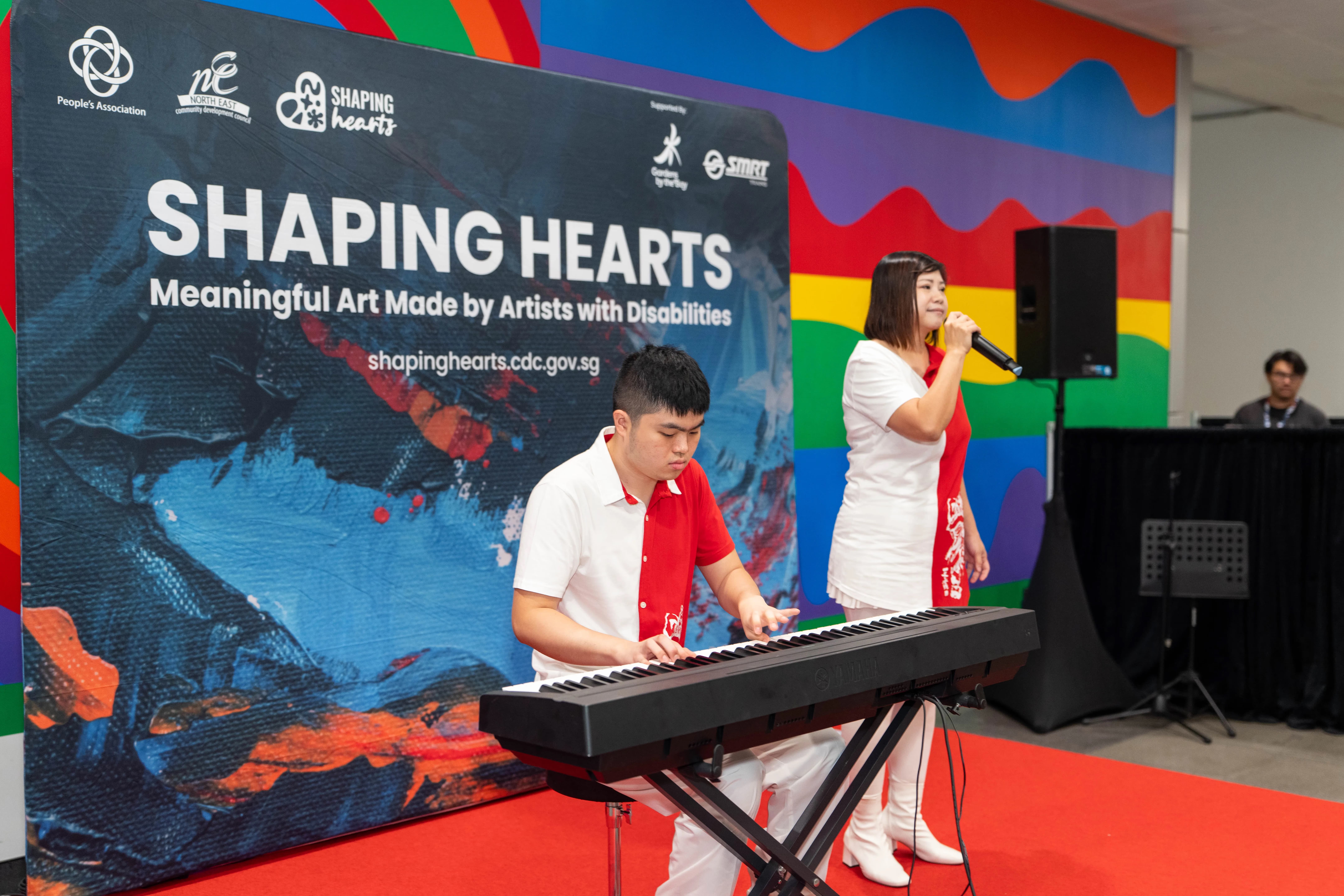 Performers on stage performing in front of a backdrop displaying “Shaping Hearts.”